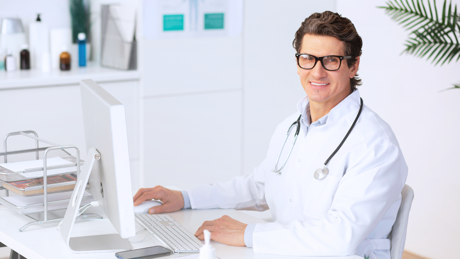 A man in a white coat, using his expertise, sits at a desk, working on a computer in a professional setting.