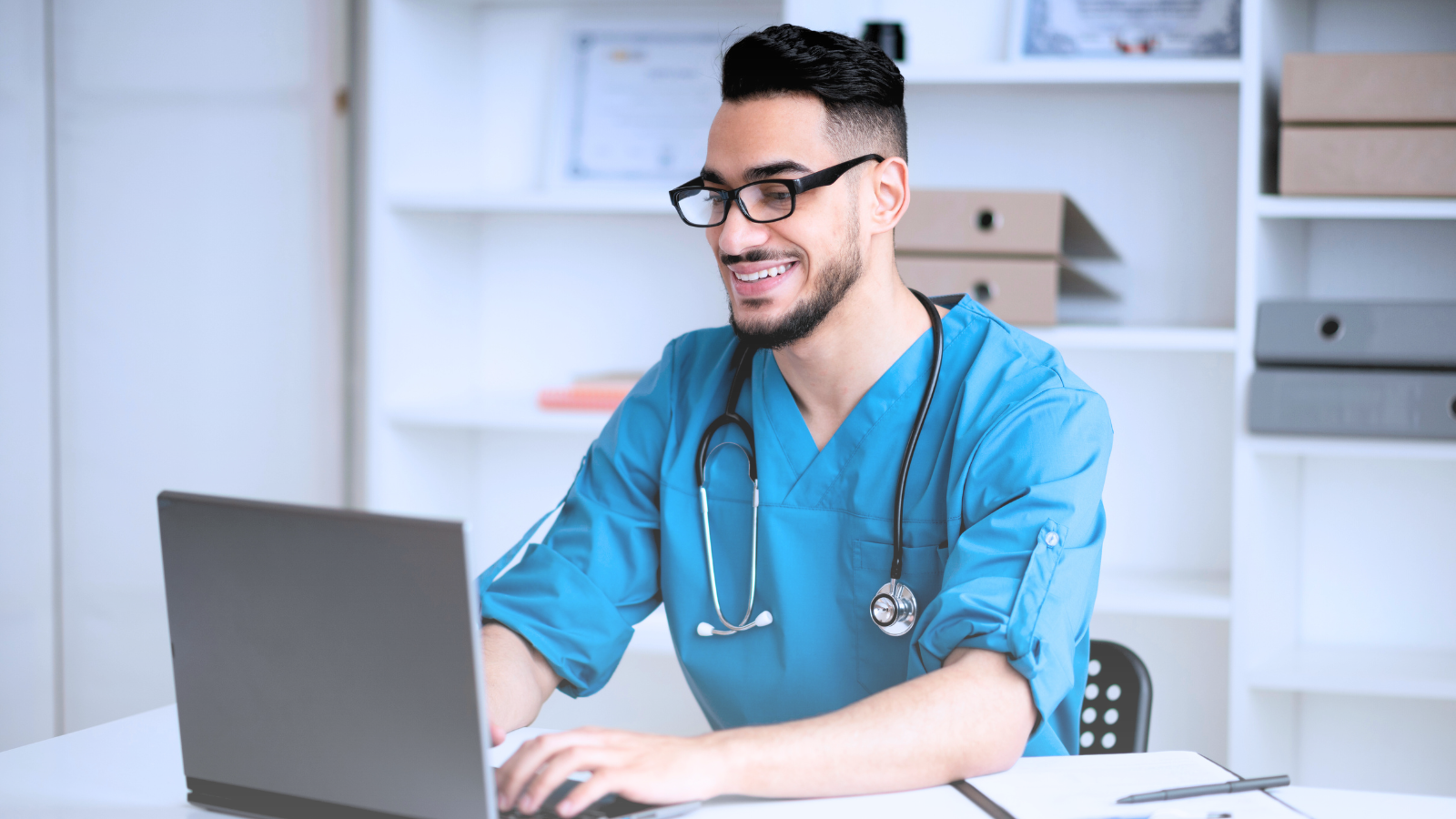 A smiling male doctor in a blue shirt works on a laptop, conveying a sense of expertise, professionalism, and approachability.