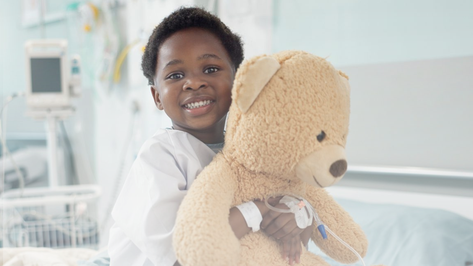 A young boy in a hospital room holds a teddy bear, looking comforted and hopeful amidst the medical environment.