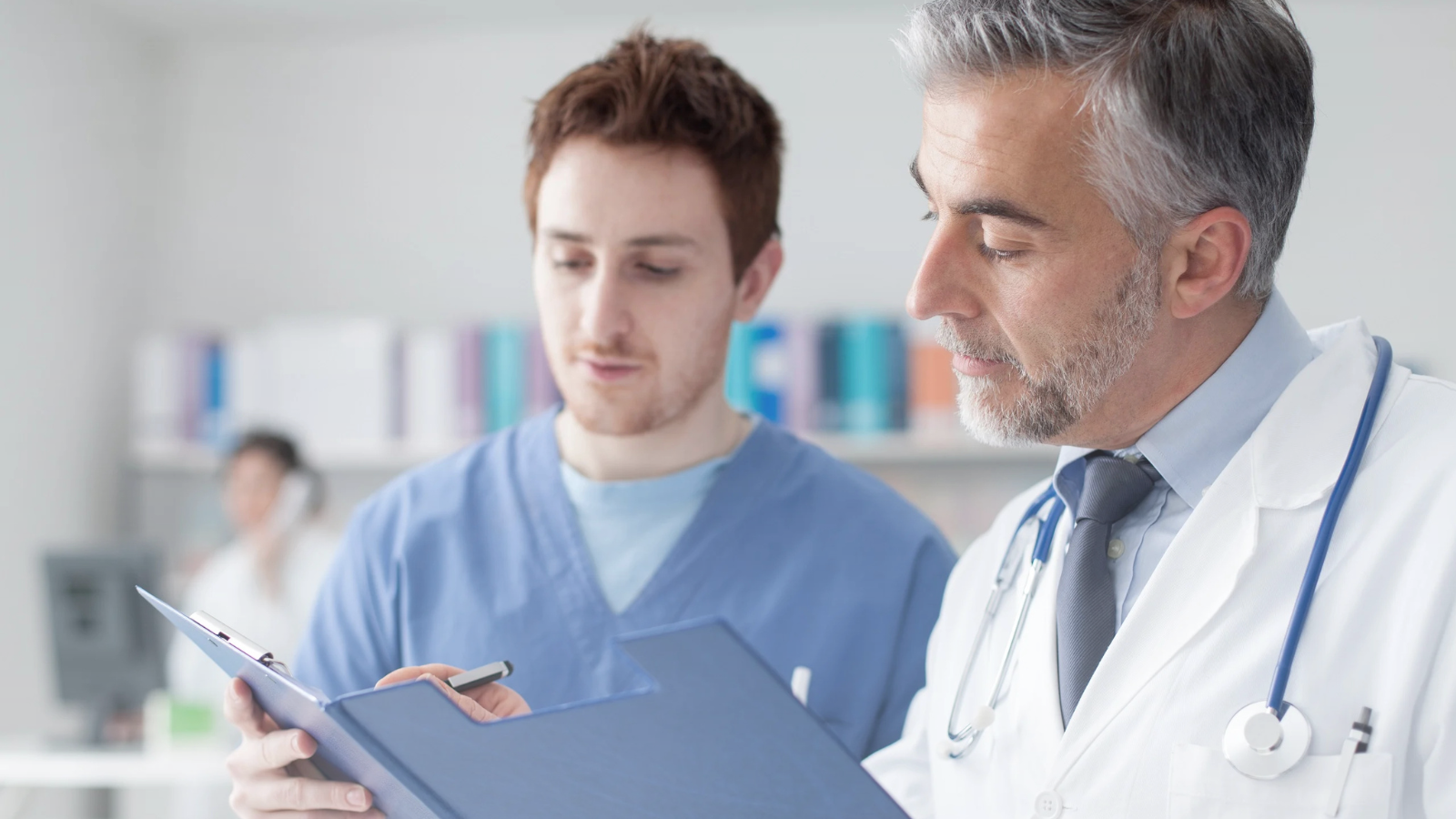 A male doctor and a male nurse review a clipboard together in a medical setting.