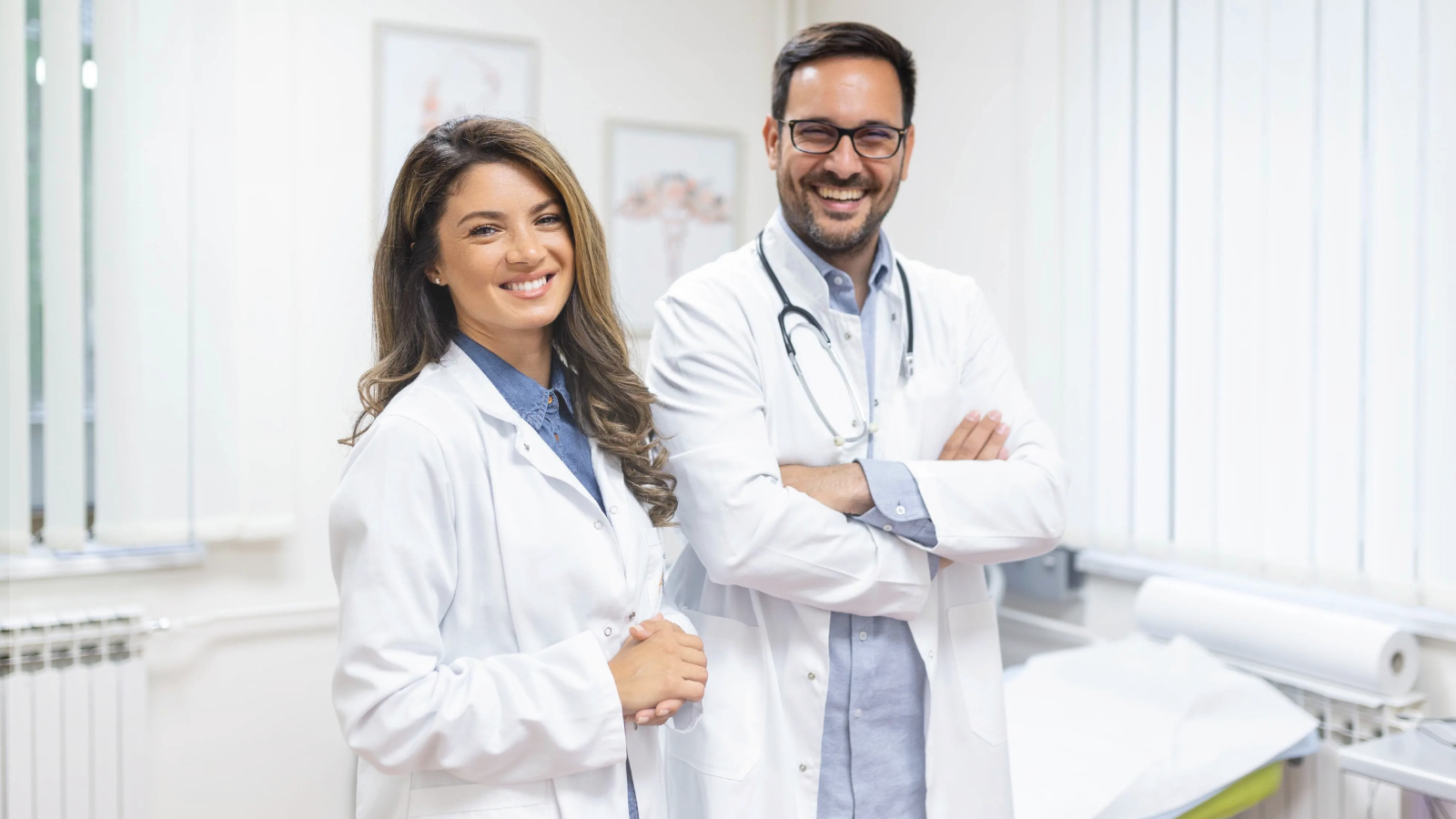 A man and woman doctors in white lab coats stand beside a hospital bed, discussing patient care.
