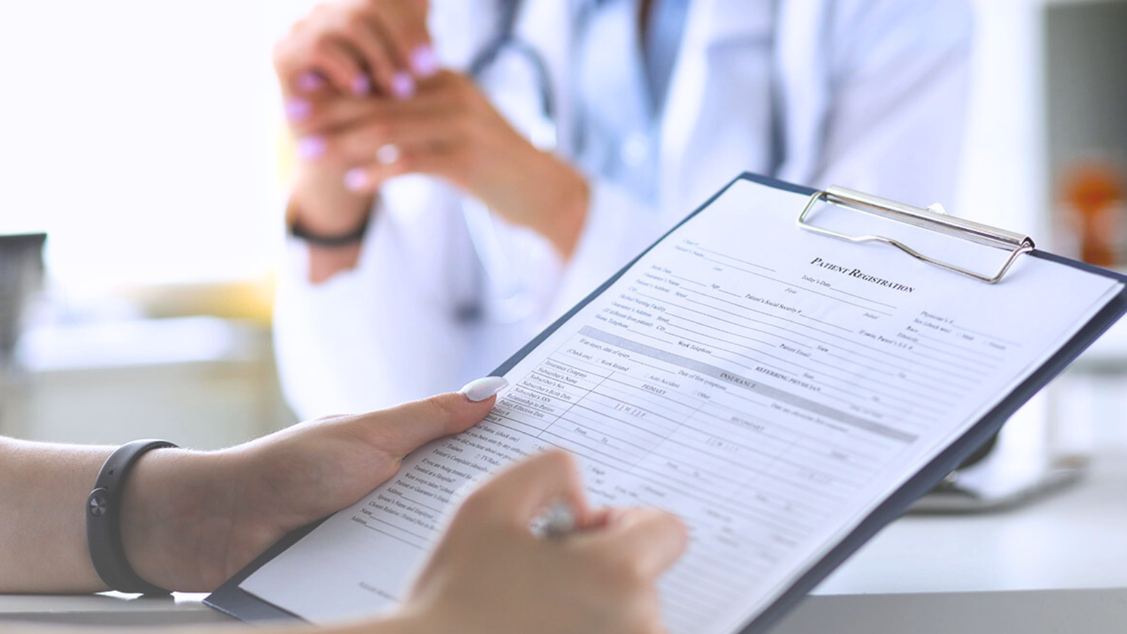 A patient holds a clipboard while a doctor sits across from them in a medical office setting.